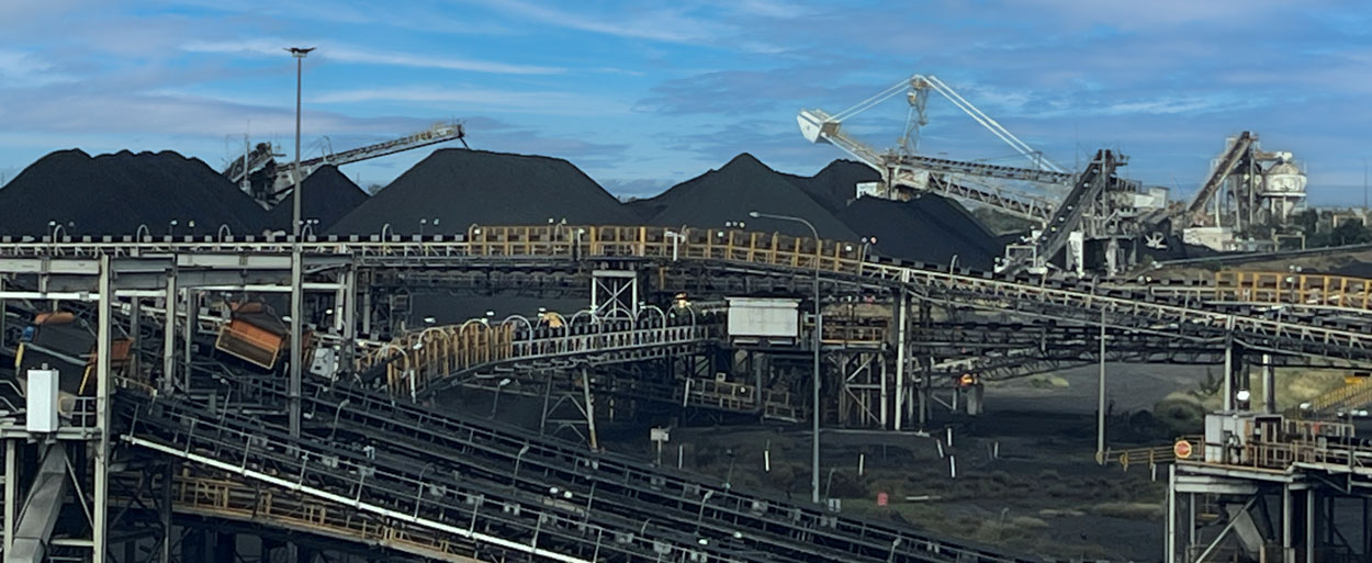 Coal handling and processing facility with multiple conveyors, stockpiles of coal, and stacking-reclaiming machinery operating under a blue sky